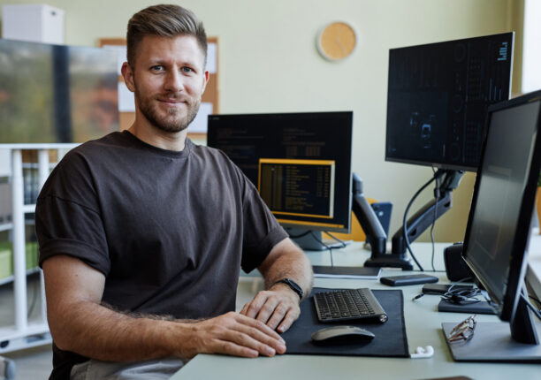 Portrait Of Computer Programmer at Workplace Portrait of smiling software developer looking at camera while sitting at desk with computers